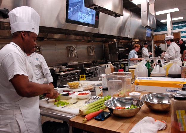 Eine Gruppe von Köchen in einer Küche, mit einem im Vordergrund in einer weißen Schürze und Mütze stehend, der ein Messer hält und von verschiedenen Utensilien, Schüsseln, Gemüse, einem Handy und Flaschen umgeben ist, mit einem Fernseher und Deckenlampen im Hintergrund.