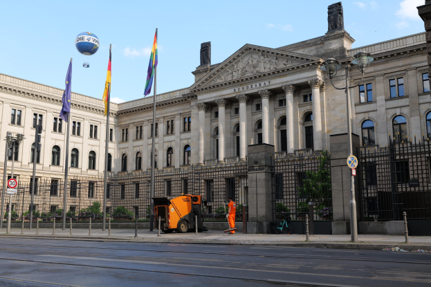 Großes modernes Gebäude mit vielen Fenstern, als Bundestag in Berlin identifiziert, umgeben von Straßeninfrastruktur und Fußgängern, mit Flaggen und Bäumen im Vordergrund und einem bewölkten Himmel im Hintergrund.