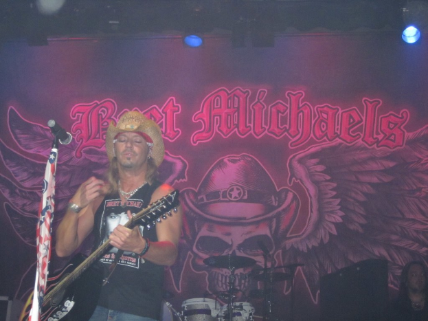 Eine Musik-Konzertszene mit Scheinwerfern oben, einem Banner im Hintergrund, Drogen in der Mitte unten, einer sitzenden Person in der rechten unteren Ecke und einem stehenden Gitarristen mit Mikrofon vorne.
