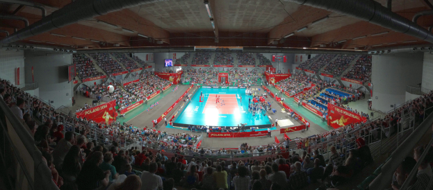 Eine große Halle mit Zuschauern bei einem Volleyballspiel, mit Bannern, Bildschirmen, Deckenlampen und Geländern unten.
