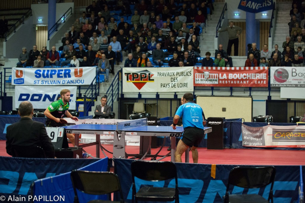 Zwei Männer spielen Tischtennis in einer Halle, umgeben von Zuschauern, leeren Stühlen und Werbetafeln, mit Text in der unteren linken Ecke.