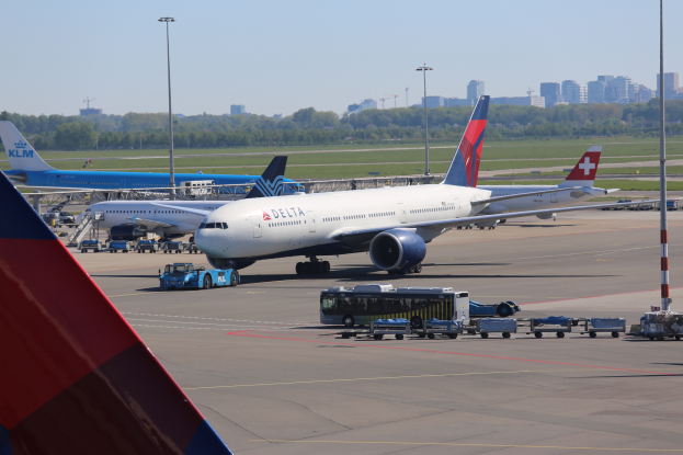 Ein Boeing 777-300ER der Delta Air Lines auf dem Frankfurter Flughafen, umgeben von Fahrzeugen und Laternenmasten, sowie Gebäuden, Bäumen und einem klaren blauen Himmel im Hintergrund.