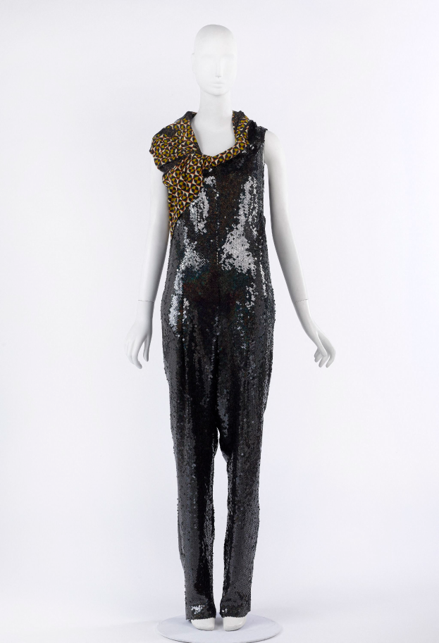 Ein Schaufensterpuppen-Mann in einem schwarzen und goldenen Pailletten-Jumpsuit mit einem Leoparden-Muster-Schal um den Hals, die Pailletten funkeln und reflektieren das Licht.
