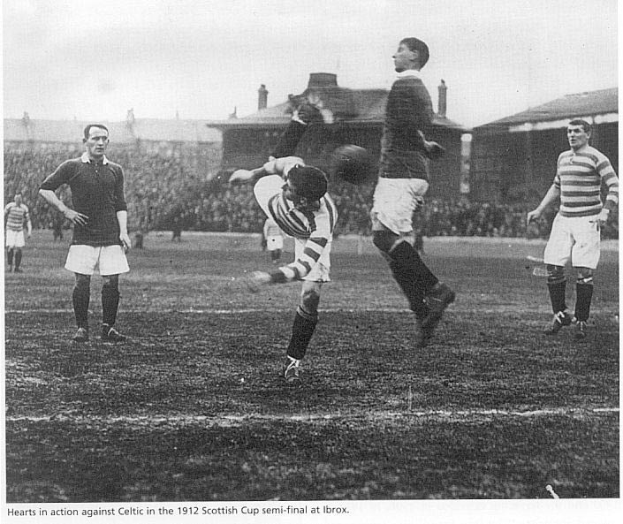 Eine Gruppe von Männern beim Fußballspielen auf einem Feld mit Zuschauern auf den Tribünen und Gebäuden im Hintergrund während des Halbfinals des schottischen Pokals 1912 im Ibrox.