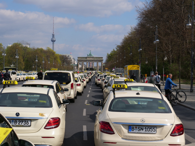 Eine lange Reihe von Taxis, die an einer belebten Straße in Berlin, Deutschland, geparkt sind, mit Fahrradfahrern und Fußgängern auf dem Bürgersteig, flankiert von Laternenpfählen und Bäumen, und ein Bogen und ein Turm im Hintergrund unter einem bewölkten Himmel.