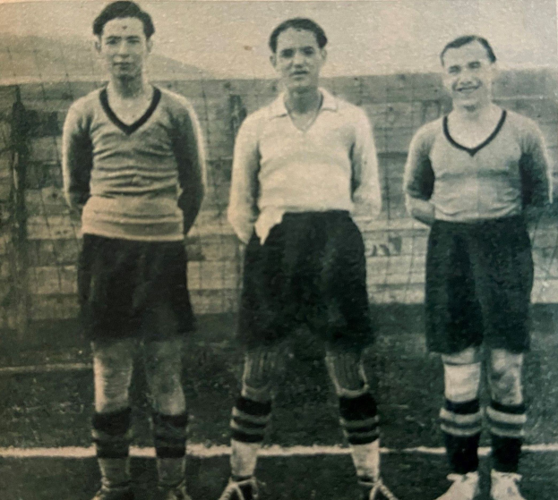 Drei Männer in Fußballtrikots und Fußballschuhen stehen Seite an Seite auf einem Fußballfeld mit einer Wand im Hintergrund.