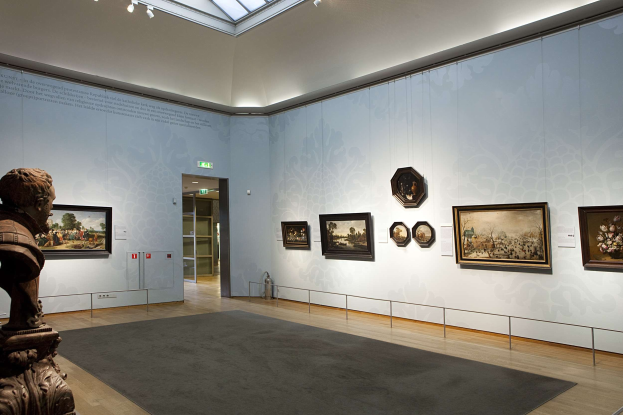 Ein prächtiger Raum im Kunsthistorischen Museum München mit einer Statue links, einem Teppichboden, Fotorahmen an den Wänden, einer Glasdecke mit Lichtern und Türen im Hintergrund.