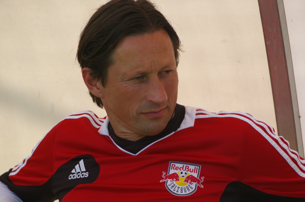 Ein Mann in einem roten und schwarzen T-Shirt sitzt auf einer Bank vor einer Wand und hat einen selbstbewussten und entschlossenen Gesichtsausdruck, der ihn wie einen professionellen Fussballspieler aussehen lässt.