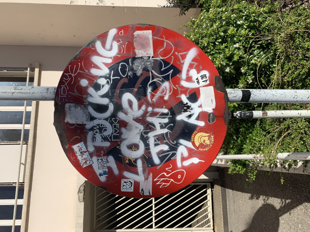 Ein rotes Schild mit Graffiti auf einem Pfahl vor einem Gebäude mit Fenstern und Bäumen im Hintergrund.