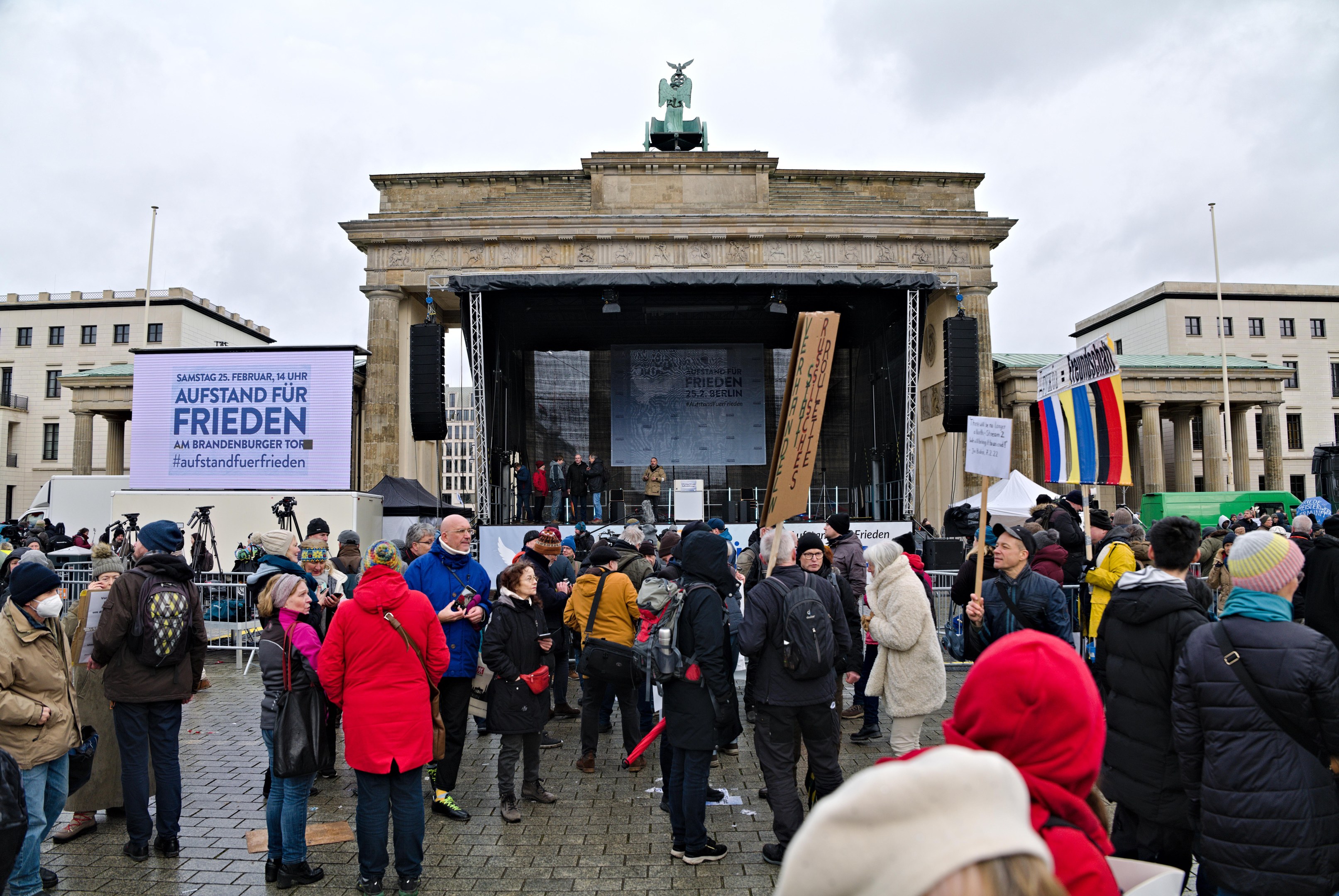 Eine Menschenmenge steht vor einem Gebäude mit einer Bühne, auf der Redner und ein Bildschirm zu sehen sind, umgeben von Fahnen und Transparenten mit Texten, während einer Demonstration in Berlin.