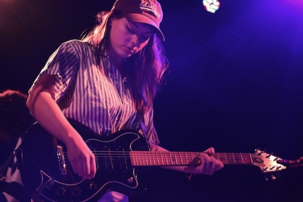 Eine Frau mit Mütze spielt Gitarre auf der Bühne bei einem Musikkonzert, während zahlreiche Lichter den Hintergrund erhellen.