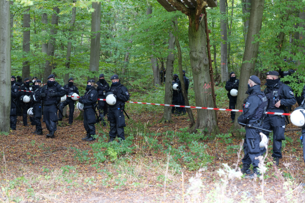 Eine Gruppe von Polizisten in Helmen und mit Waffen in der Hand steht in einem bewaldeten Gebiet mit einem Schuppen im Hintergrund und einem Band an einem Pfahl im Vordergrund.