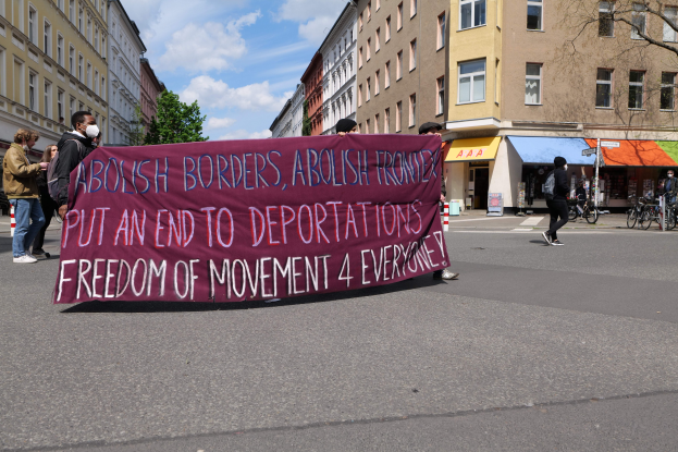Eine Gruppe von Menschen marschiert mit einem Banner, auf dem "Abolish Borders, Abolish Frontiers, Put an End to Deportations, Freedom of Movement 4 Everyone" steht, durch eine Straße mit Gebäuden, Bäumen, Fahrrädern und einer bewölkten Himmel.