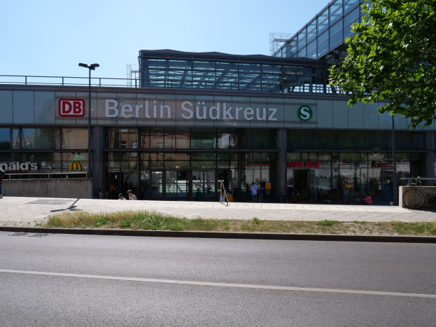 Großes gläsernes Berliner Südkreuz-Bahnhof in Deutschland mit sichtbarem Text, umgeben von Straßeninfrastruktur, Fahrzeugen, Fußgängern, Fahrrädern, Bäumen und einem klaren Himmel.
