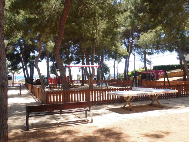 Ein Park mit einem zentralen Tischtennis-Tisch, umgeben von Bänken, einem Zaun, Schaukeln, Pfosten, Pflanzen und Bäumen, mit Blick auf einen See unter einem klaren blauen Himmel.