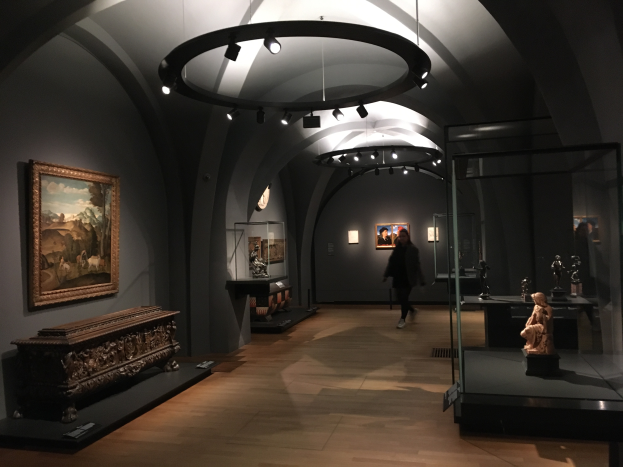 Eine Person geht durch ein Museum mit Kunst an den Wänden, einer beleuchteten Decke und einer Glasvitrine auf der rechten Seite mit Skulpturen und Gegenständen.