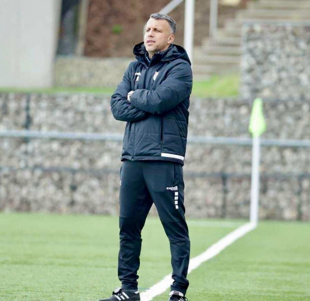 Ein Mann steht auf einem Fußballfeld mit verschränkten Armen und trägt eine schwarze Jacke und Hose, mit einem Zaun, Treppe und einer Wand im Hintergrund.