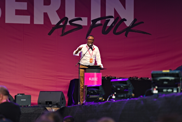 Ein Mann in einem weißen Hemd und braunen Hosen steht auf einer Bühne und hält ein Mikrofon in der Hand, mit einem pinken Banner "Berlin as Fuck" im Hintergrund und einer Menge davor.