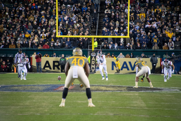 Fußballspieler in Sportkleidung und Helmen konkurrieren auf einem Feld mit Zuschauern im Hintergrund, Stadionstrukturen und sichtbaren Schildern.