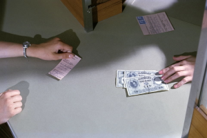 Eine Person gibt Geld an einer Kasse an eine andere Person weiter, mit Geldscheinen und Papieren auf dem Tisch und einer Tür rechts daneben.