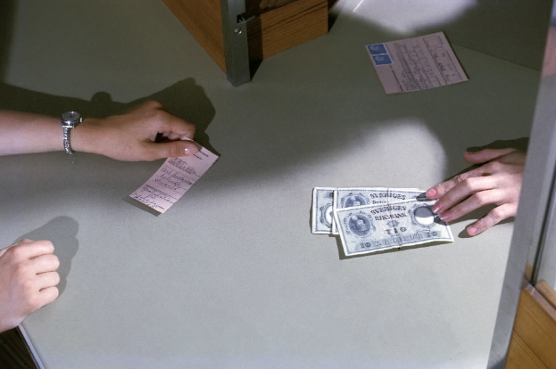 Eine Person gibt Geld an einer Kasse an eine andere Person weiter, mit Geldscheinen und Papieren auf dem Tisch und einer Tür rechts daneben.
