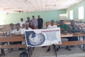 Eine Gruppe von Menschen sitzt an Tischen in einem Klassenraum mit grünen Wänden und Fenstern auf beiden Seiten und hält ein Banner in die Höhe, auf dem "Wipeda Awareness and Training at Ramama Polytechnic Maiduguri" steht, mit Taschen auf dem Boden und ein paar Menschen im Hintergrund.