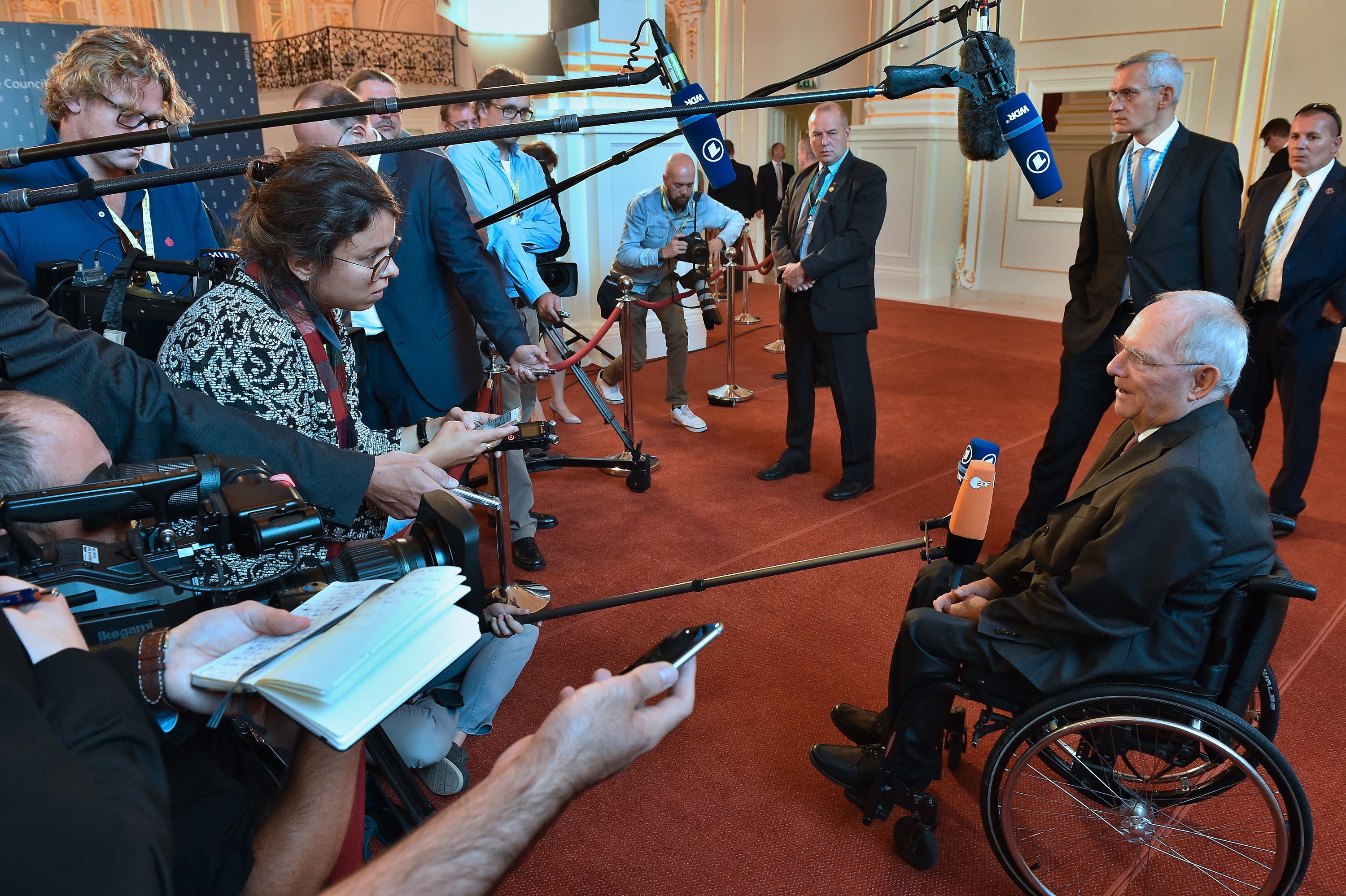 Ein Mann im Rollstuhl in Anzug und Krawatte von Reportern mit Kameras und Mikrofonen umgeben