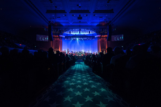 Ein großer Auditorium mit sitzenden Zuschauern, eine Bühne mit Musikern und Überkopfbeleuchtung und Fernseher, was auf einen Konzertsaal während der Nationalen Convention 2019 hindeutet.