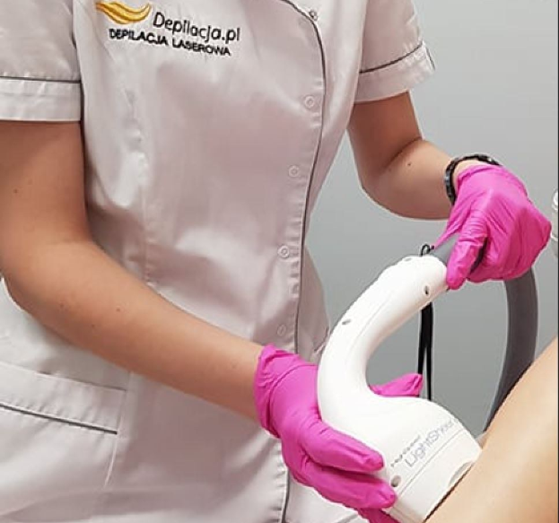 Eine Frau in einem weißen Hemd und pinken Handschuhen bedient eine weiße Laser-Haarentfernungsmaschine an ihrem Arm, während sie an einer Wand steht.