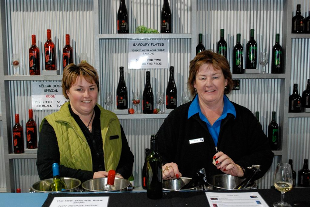 Frauen an einem Tisch mit einem Glas, einer Flasche und Papieren mit Text, vor Regalen mit Flaschen, Gläsern und Text an der Wand.