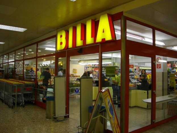 Ein Markt mit einem Eingang in der Mitte, Mülltonnen daneben, eine Reihe von Einkaufswagen auf der linken Seite, Deckenbeleuchtung und einige Menschen im Inneren.