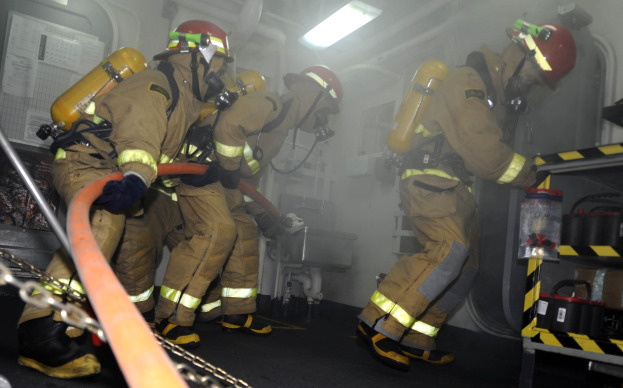 Feuerwehrleute in Schutzausrüstung, einschließlich Helmen und Sauerstoffflaschen, arbeiten an einem Feuerwehrauto, mit einem Gestell voller verschiedener Gegenstände auf der rechten Seite und Rohren, Lampen und anderen Gegenständen im Hintergrund.