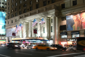 Eine nächtliche Stadtstraße mit fahrenden Fahrzeugen, ein Gebäude mit Fenstern, Säulen und Fahnenmasten, beleuchtete Schilder mit Text und einem Frauenbild, sowie helle Straßenbeleuchtung.