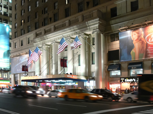 Eine nächtliche Stadtstraße mit fahrenden Fahrzeugen, ein Gebäude mit Fenstern, Säulen und Fahnenmasten, beleuchtete Schilder mit Text und einem Frauenbild, sowie helle Straßenbeleuchtung.
