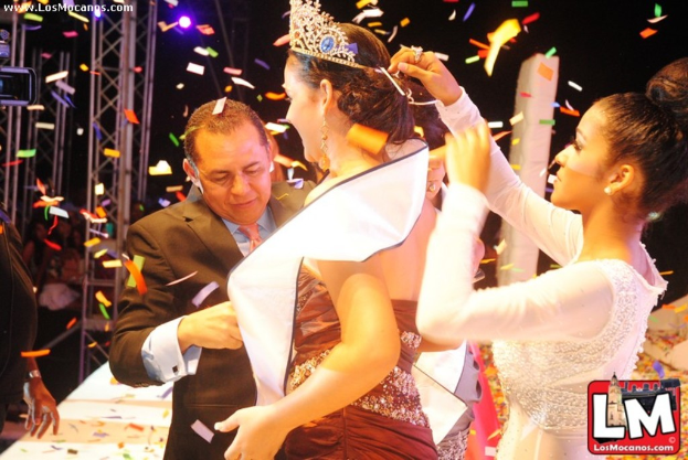 Ein Mädchen auf der Bühne mit einer Krone, während ein anderer Mädchen sie hinter ihr anpasst und ein Mann in einem schwarzen Anzug ihr die Hand schüttelt, alles vor einem dunklen Hintergrund mit Metallrahmen.