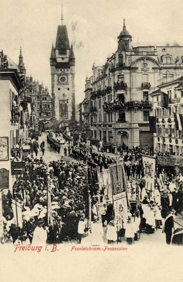 Ein Schwarz-Weiß-Foto einer belebten Straße in Freiburg, Deutschland, mit Menschen, die gehen, Gebäuden auf beiden Seiten und einem Uhrturm im Hintergrund, als alte Postkarte aus den frühen 1900er-Jahren bezeichnet.