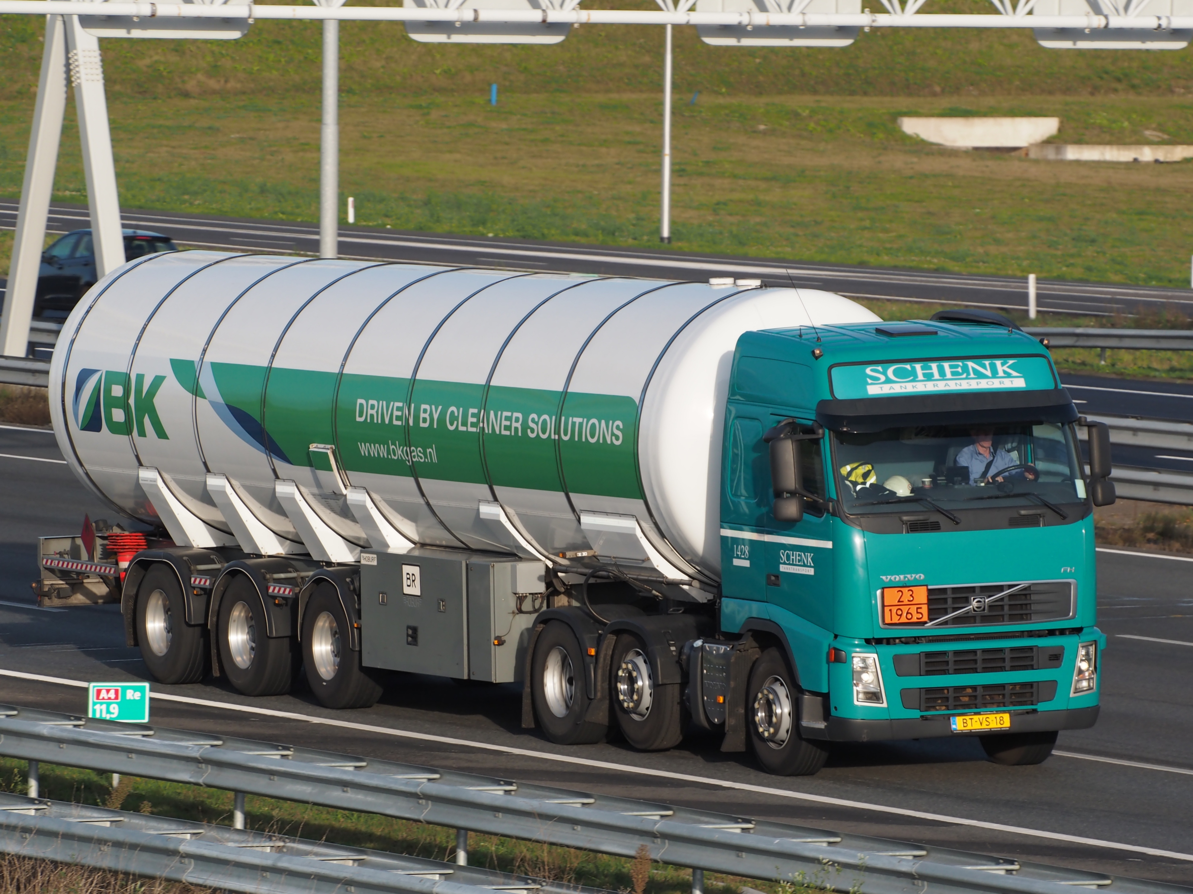 Ein großer Tanklastzug fährt auf einer Autobahn mit einer Person am Steuer, umgeben von Leitplanken und Gras, mit Polen und Schildern mit Wörtern im Hintergrund.