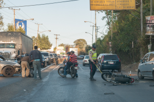 Eine Gruppe von Menschen steht um ein verunglücktes Motorrad auf der Seite einer Straße mit mehreren Fahrzeugen, darunter ein Lastwagen, und einer Hintergrund von Bäumen, Polen, Lampen, Tafeln und Himmel.