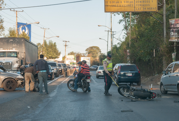 Eine Gruppe von Menschen steht um ein verunglücktes Motorrad auf der Seite einer Straße mit mehreren Fahrzeugen, darunter ein Lastwagen, und einer Hintergrund von Bäumen, Polen, Lampen, Tafeln und Himmel.