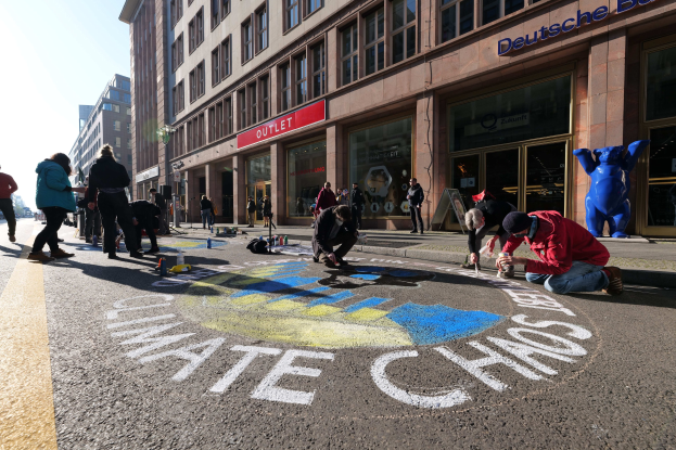Menschen sitzen vor einem Gebäude während einer Klimawandel-Demonstration in Berlin, umgeben von Flaschen und anderen Gegenständen, mit Bäumen und einem klaren blauen Himmel im Hintergrund.