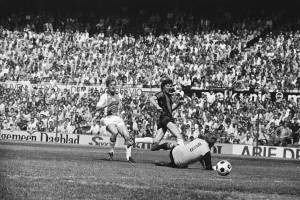 Schwarz-weißes Foto von Männern, die auf einem Feld Fußball spielen, mit Zuschauern im Hintergrund und einer 'Bundesliga 1978-79 Ajax 1-1 Hannover 96'-Fahne.