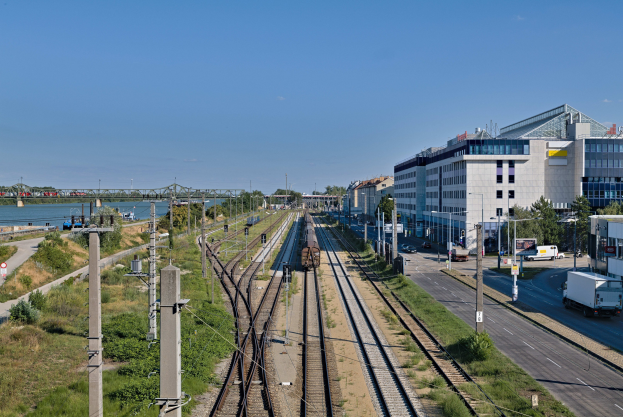 Ein Zug fährt auf Schienen neben einer Stadtlandschaft mit Gebäuden, Fahrzeugen, Straßeninfrastruktur, Grünflächen, Wasser, einer Brücke und einer bewölkten Himmel.