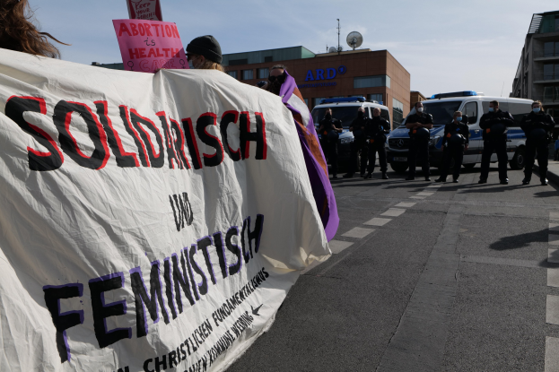 Gruppe von Menschen marschiert die Straße entlang und hält eine 'Solidarität und Feminismus'-Fahne, mit geparkten Fahrzeugen und Gebäuden im Hintergrund unter einem klaren blauen Himmel.