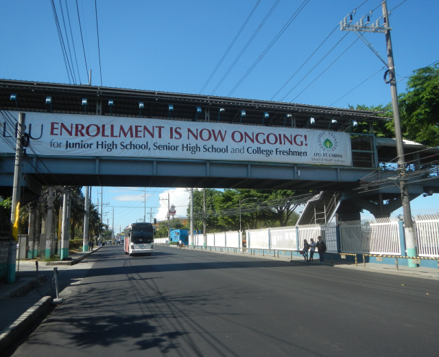 Ein Bus fährt unter einer Brücke mit einem "Jetzt Anmelden"-Schild, Fußgänger auf dem Gehweg, Strommasten mit Drähten, Bäume und ein klarer blauer Himmel im Hintergrund.