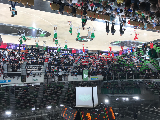 Eine Gruppe von Menschen, die Basketball in einer großen Arena spielen, mit Zuschauern in den Rängen, beleuchtet von Bildschirmen und Lichtern am unteren Bildrand.