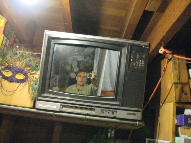 Ein Fernsehbildschirm zeigt eine Person, umgeben von Pfauenfedern, Büchern und Kartons.