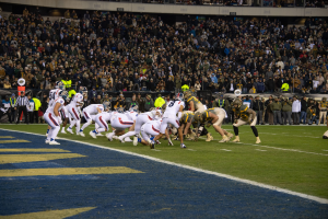 Ein Fußballspiel in vollem Gange mit Spielern auf dem Boden in Sportbekleidung und Helmen, aus der Perspektive eines Stadionplatzes.