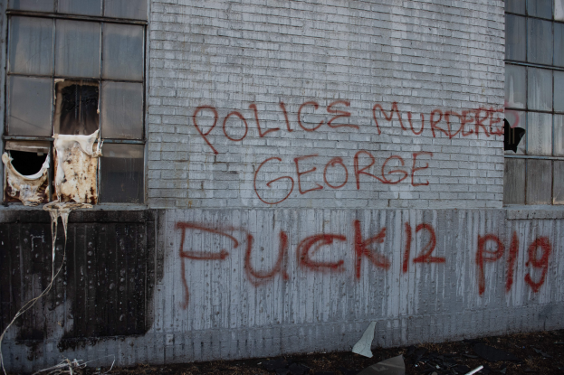 Ein Gebäude mit Graffiti, das 'Polizei ermordet George Fuck 12 P19' auf der Seite trägt, mit Fenstern und verschiedenen Gegenständen vorne auf dem Boden.