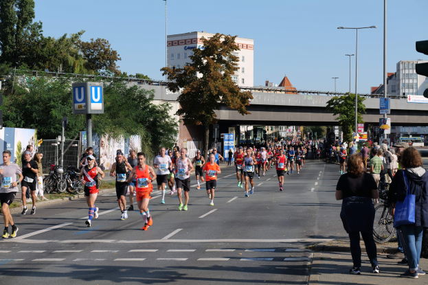 Gruppe von Menschen, die an einem Marathon auf einer von Bäumen gesäumten Straße mit Laternen, Schildern, Fahrrädern, einem Zaun, Gras, einer Brücke, Gebäuden und einem klaren blauen Himmel laufen.