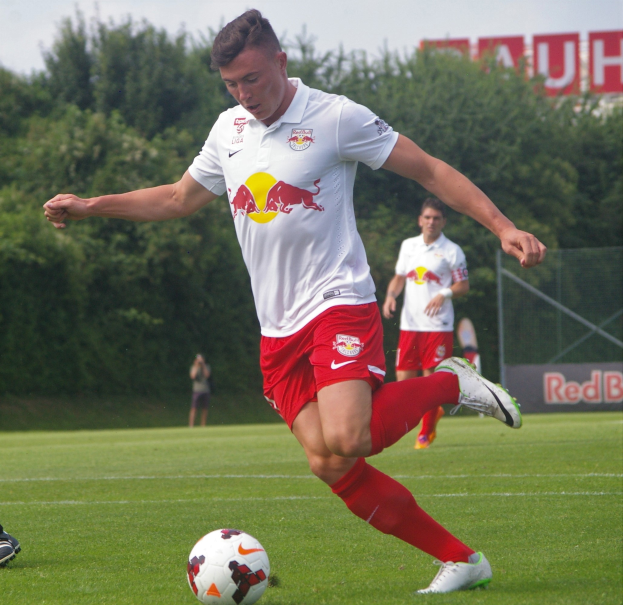 Ein Mann in einem Fußballtrikot kickt einen Ball auf einem grünen Feld mit Bäumen und einem klaren blauen Himmel im Hintergrund, wobei ein "RB Leipzig"-Schild sichtbar ist.